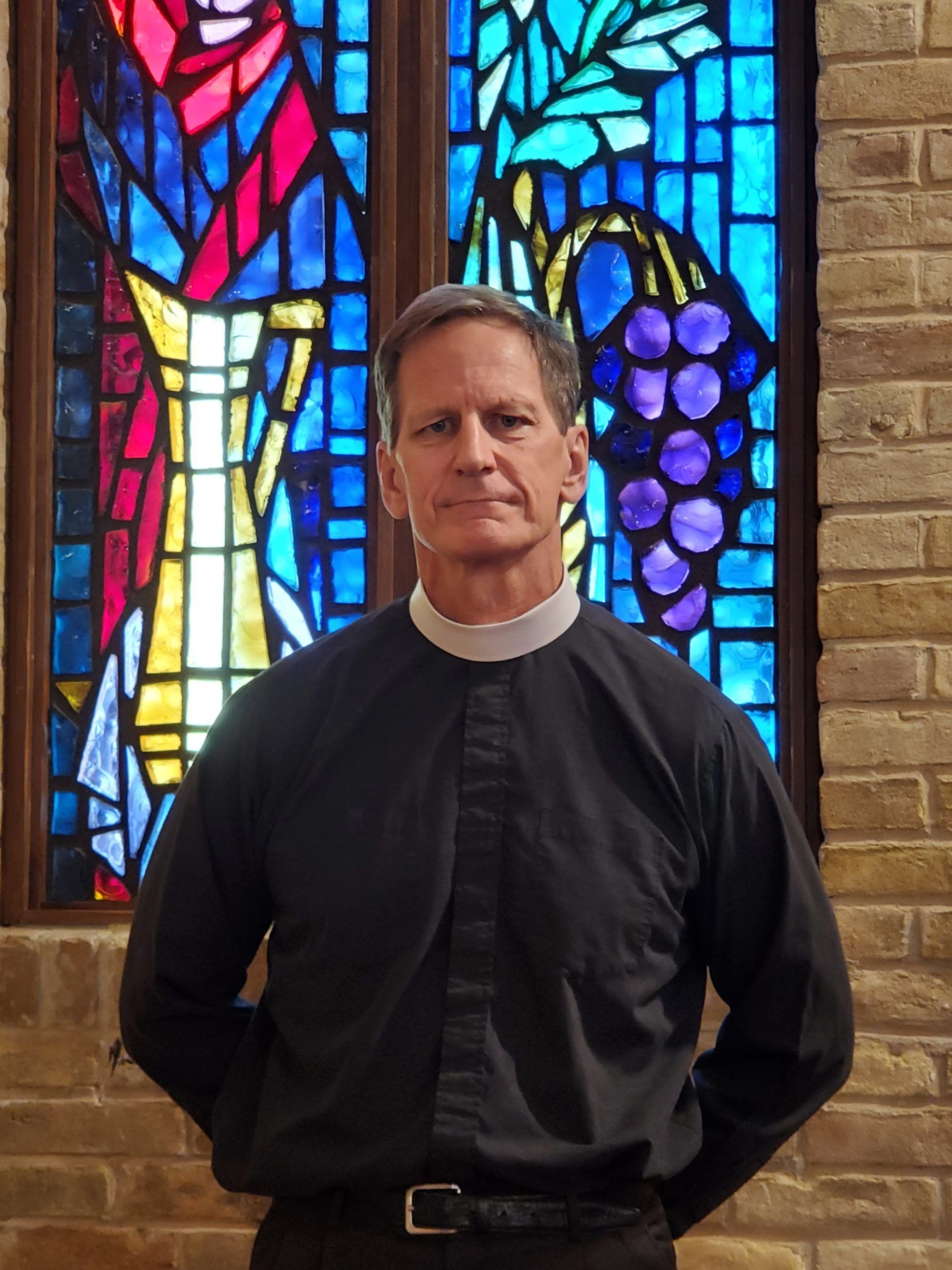 A clergy member in a black shirt and clerical collar, standing before a stained-glass window with grapes and foliage.