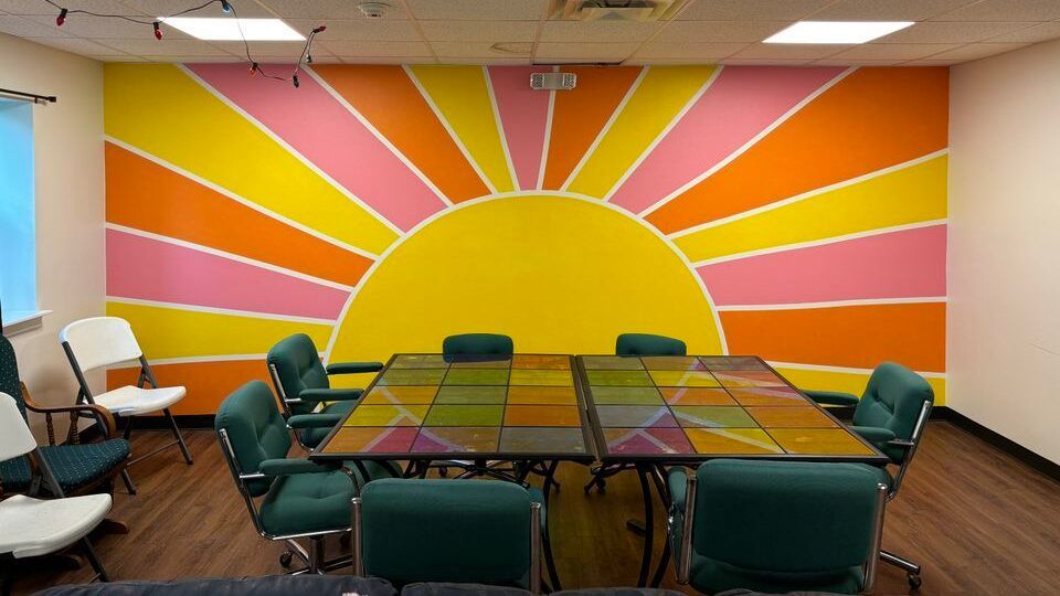 A meeting room with a large, colorful sun mural on the back wall, featuring alternating rays of yellow, pink, and orange.
