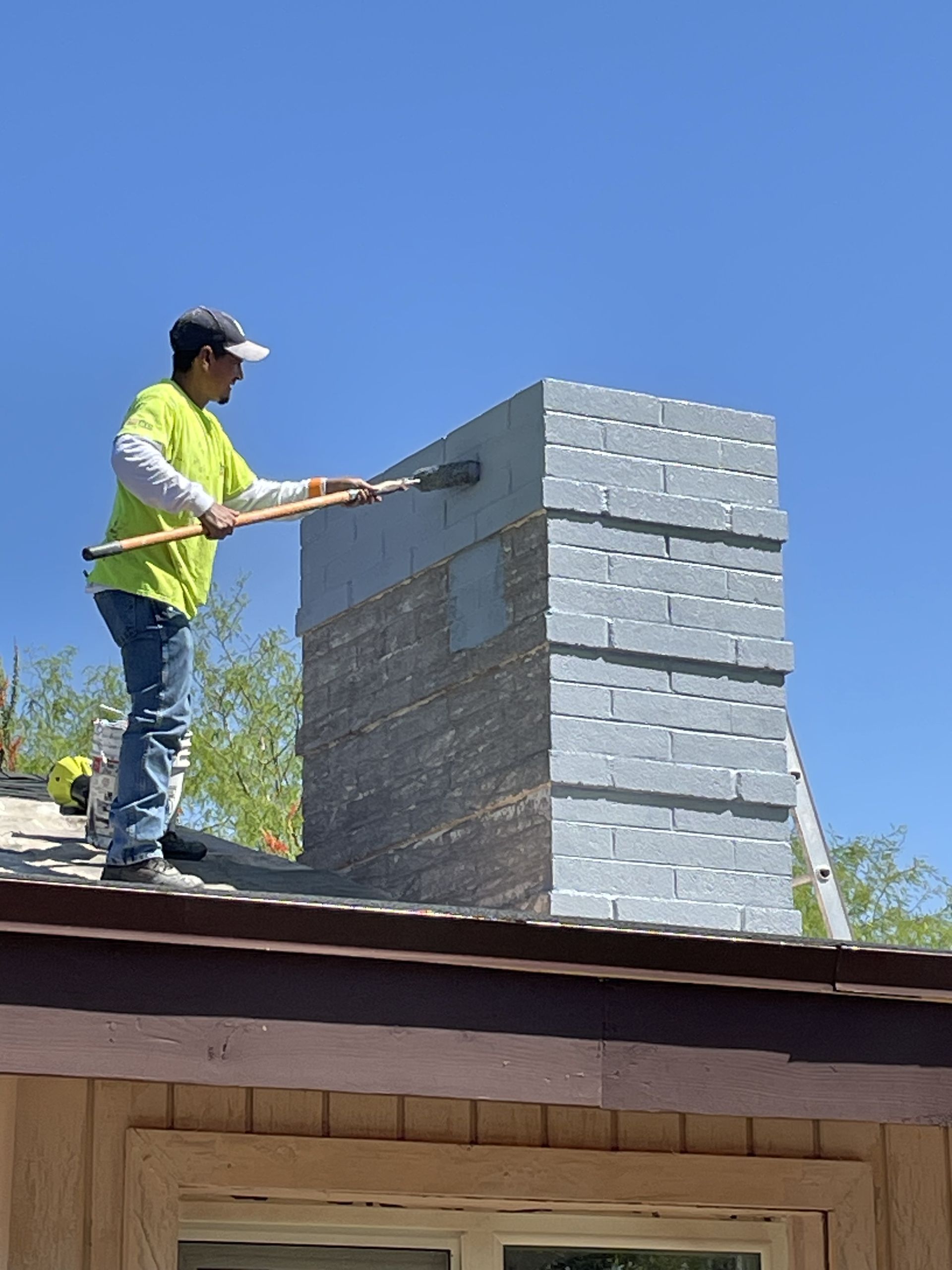 A man is standing on top of a roof painting a chimney.