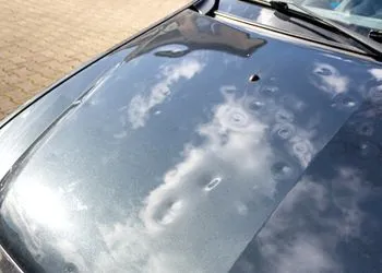Dark car hood with multiple shallow dents caused by hail, reflecting the sky on its surface.