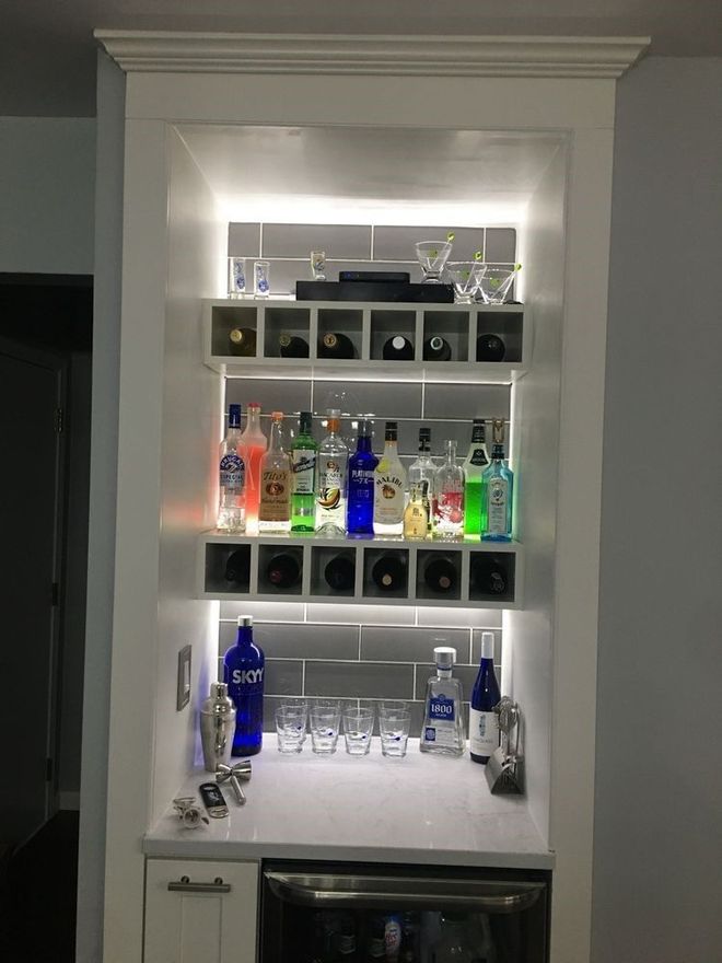 A built-in home bar with white shelving, a white countertop, lit glass tile backsplash, and various liquor bottles.
