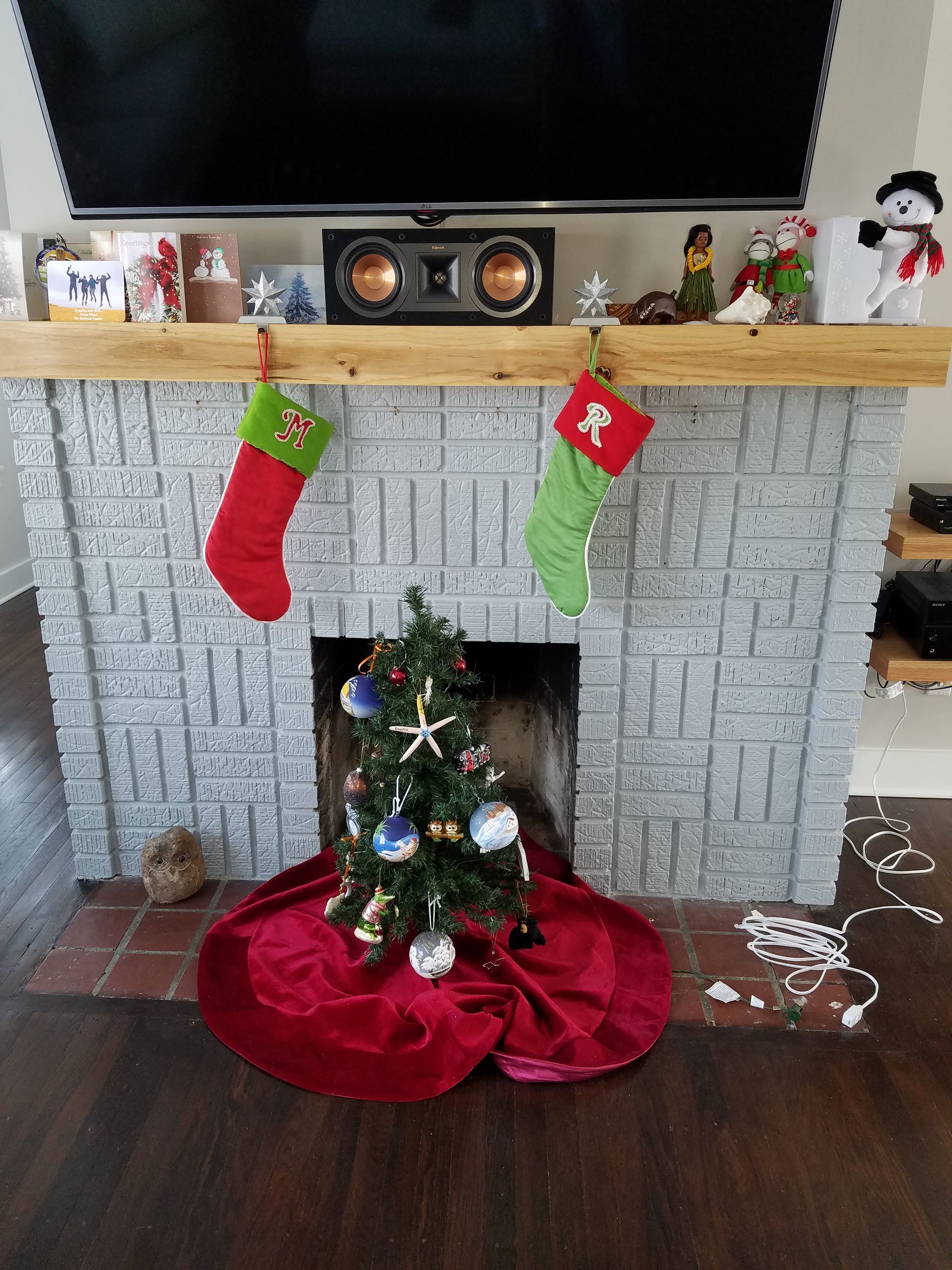 A gray brick fireplace decorated for Christmas, featuring stockings, a small tree in the hearth, and mantel figurines.