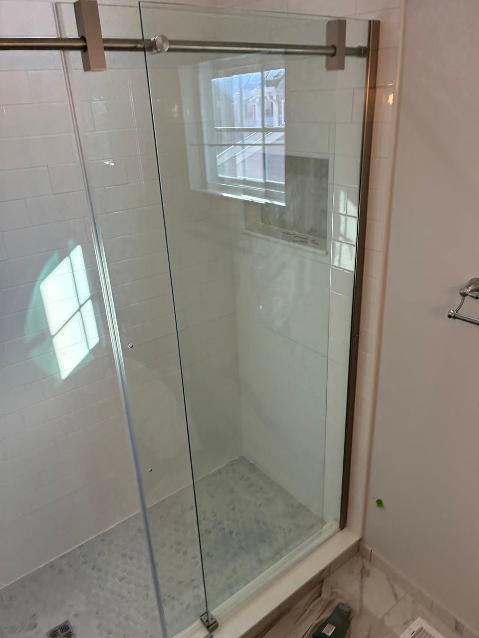 A glass sliding shower door with a metal top rail, installed in a bathroom with white tiled walls and marble flooring.