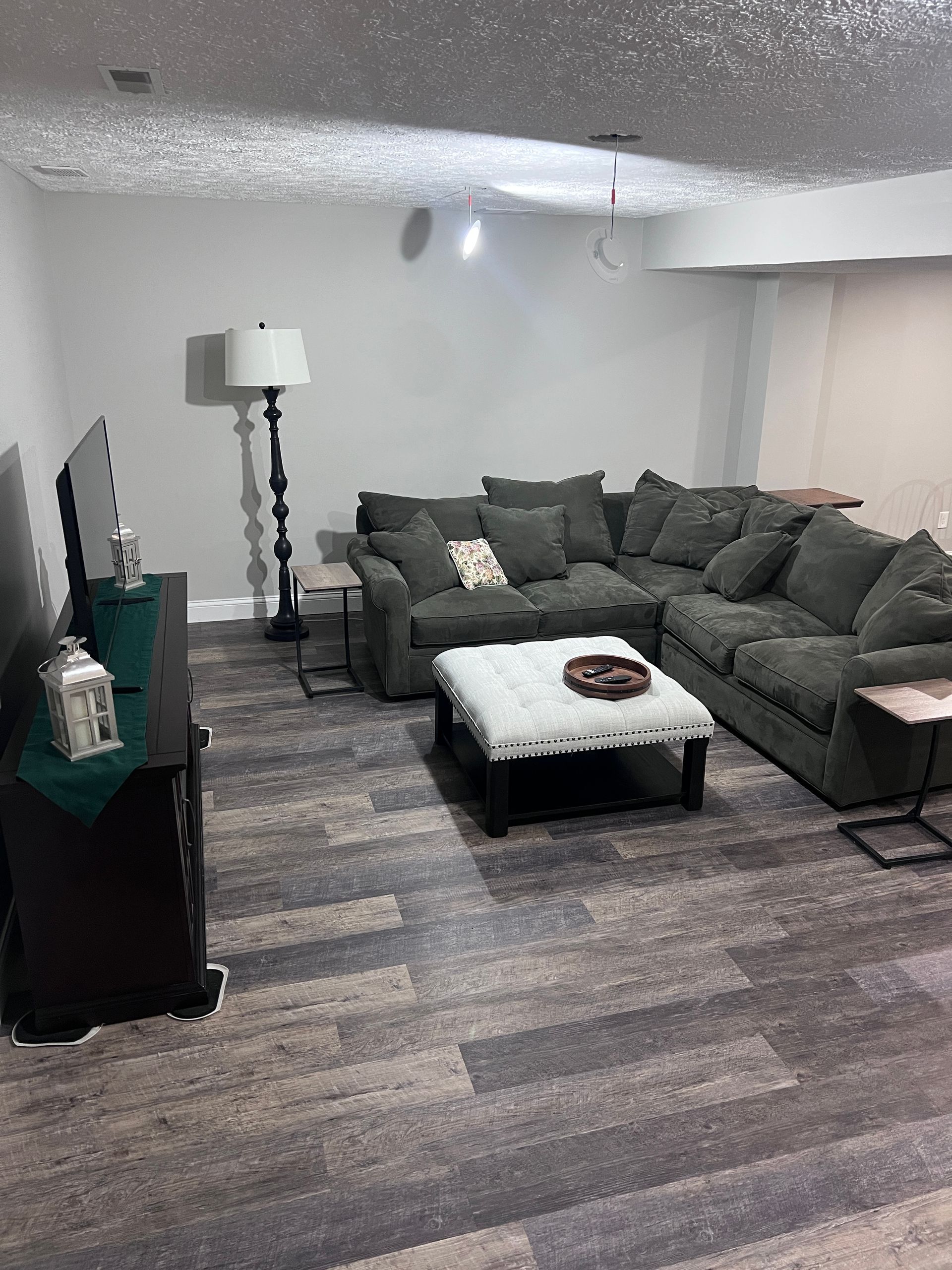 A grey L-shaped sectional sofa, white tufted ottoman, dark media console, and floor lamp in a room with patterned flooring.