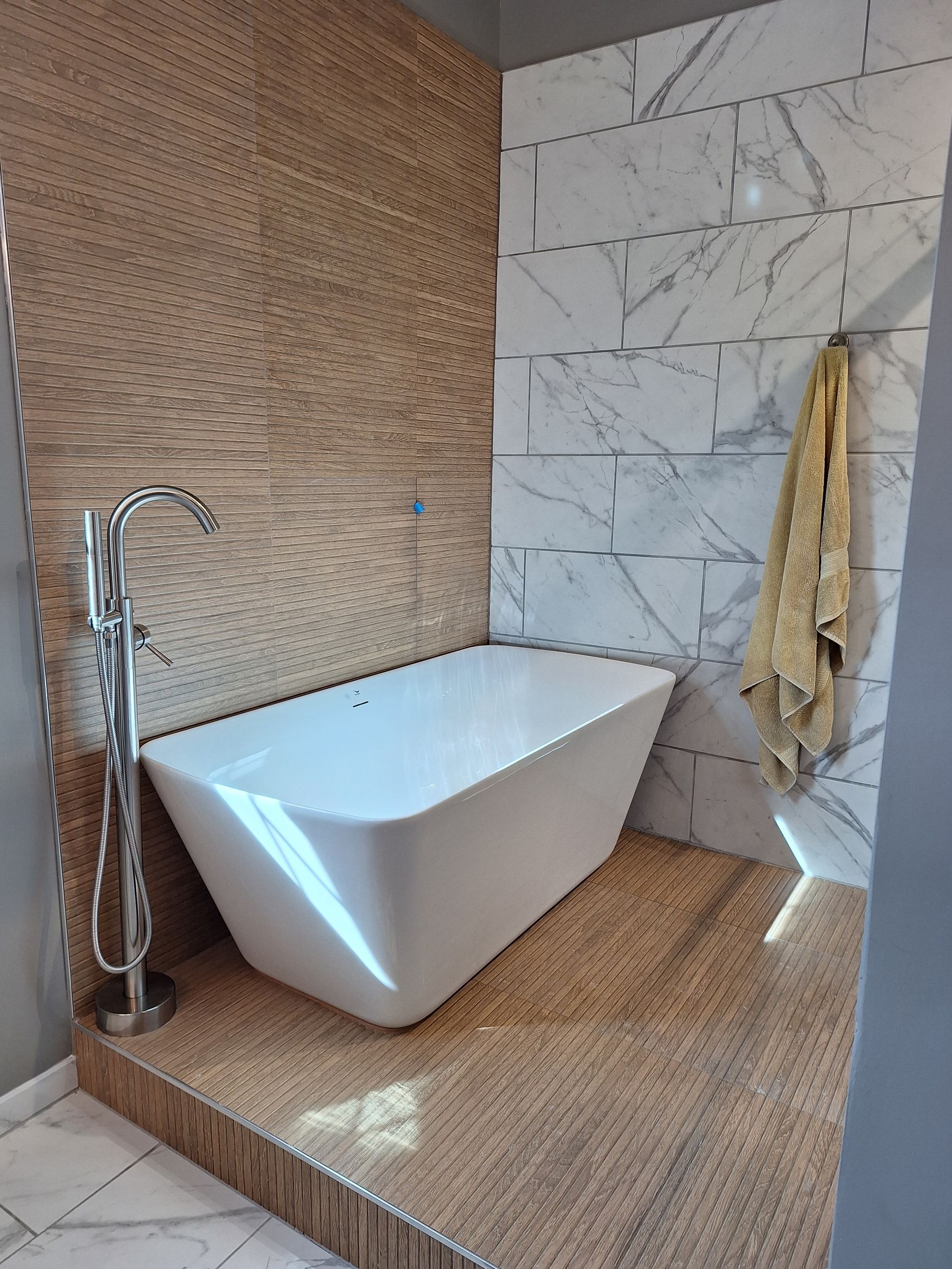 A white freestanding bathtub set on a textured, woven-style platform next to white marble wall tiles and a hanging towel.