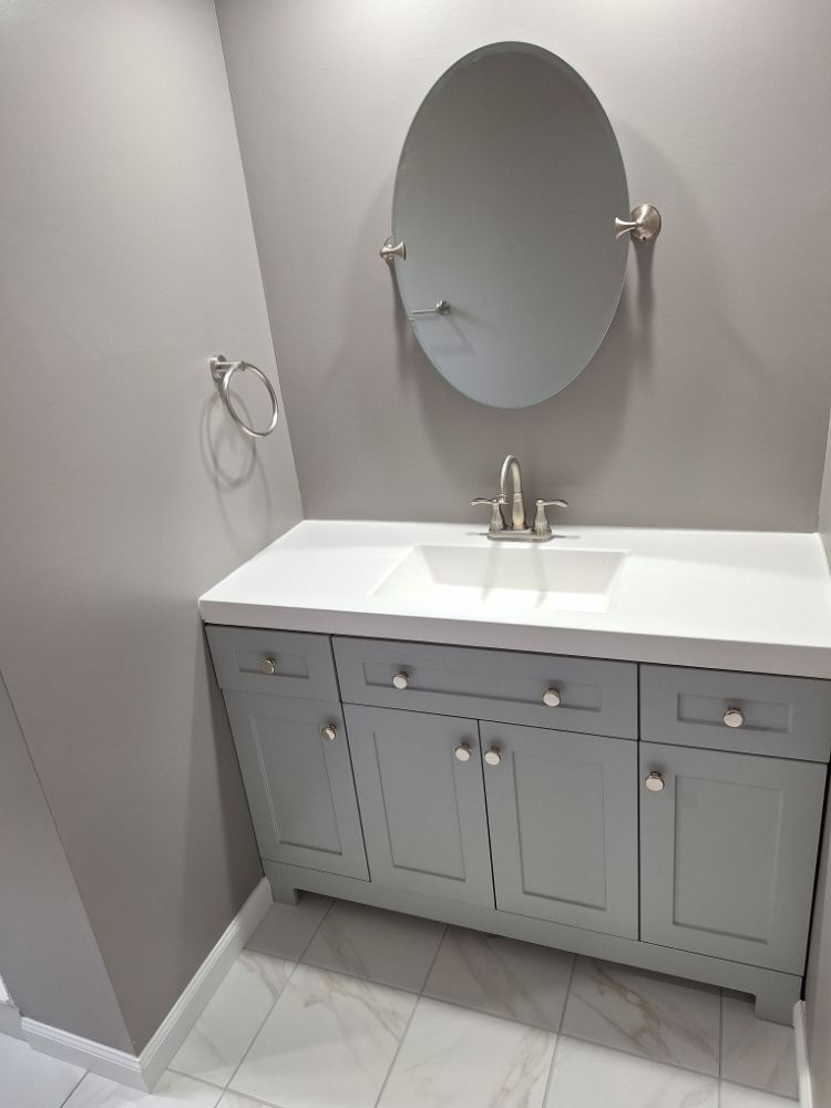 A grey bathroom vanity with a white countertop and oval mirror, featuring a chrome faucet and a towel ring on the wall.