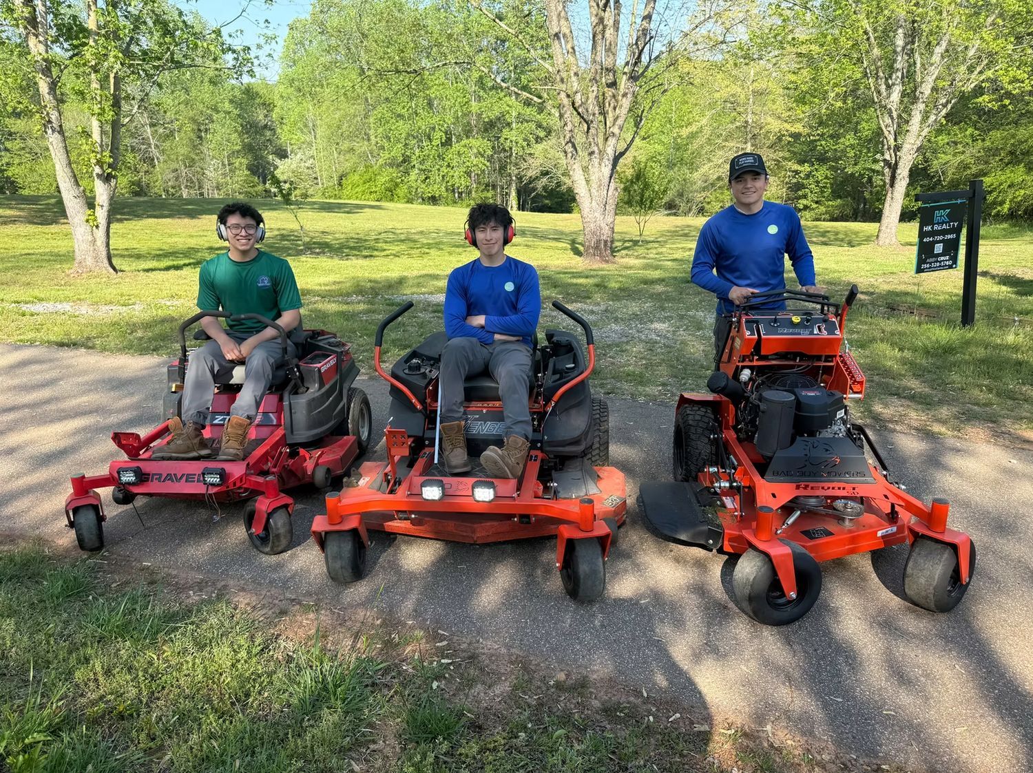 Three people with lawn mowers outdoors. Two sit on mowers; one stands behind his. Green trees in background.