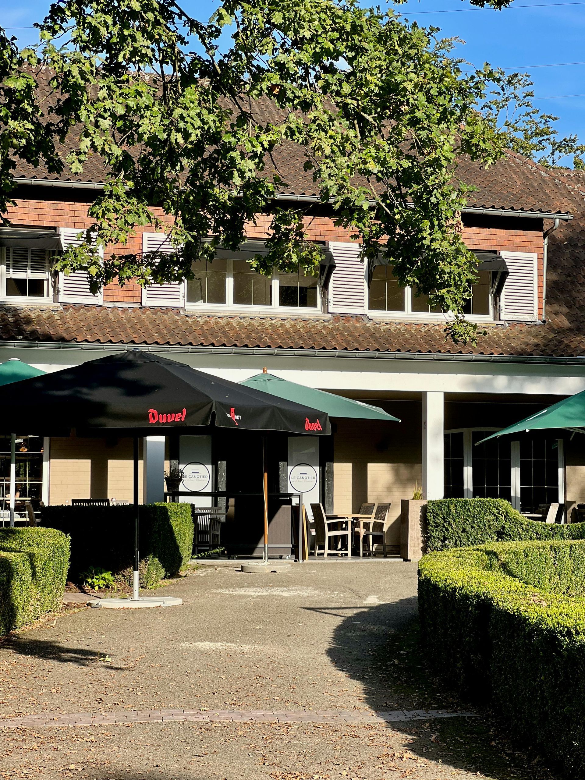 Restaurant exterior with outdoor seating under umbrellas; brick building, green hedge, trees.