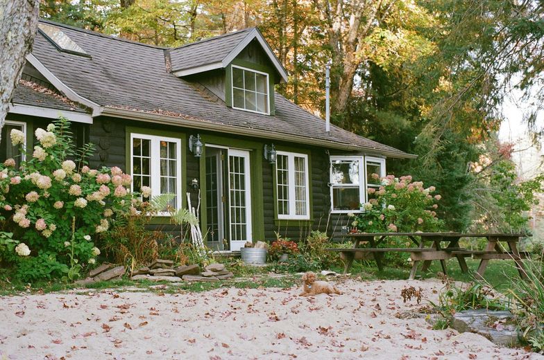 Dark green cottage with white-framed windows, surrounded by hydrangeas and fallen leaves, picnic table.