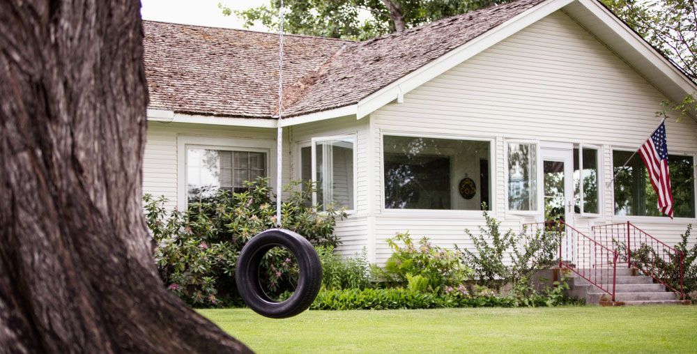 A tire swing is hanging from a tree in front of a white house.