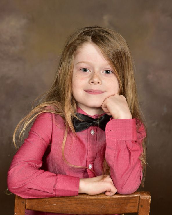 A little girl wearing a pink shirt and a black bow tie is sitting in a chair.