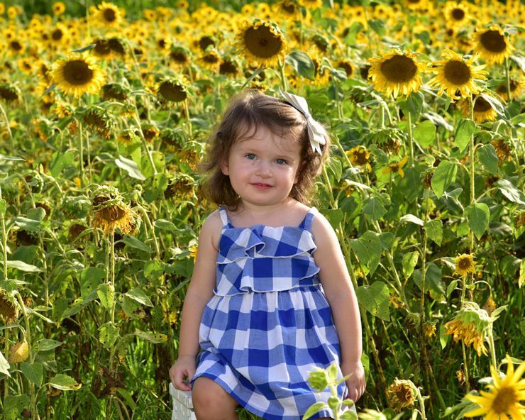 A little girl in a blue and white dress is standing in a field of sunflowers.