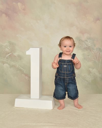 A baby in overalls is standing next to a white number one.