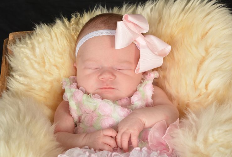 A newborn baby girl is sleeping in a basket with a pink bow on her head.