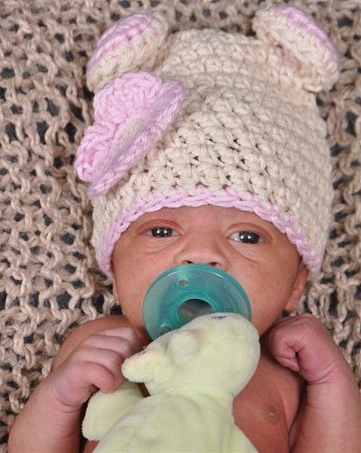 A baby wearing a crocheted hat and pacifier is holding a stuffed animal.