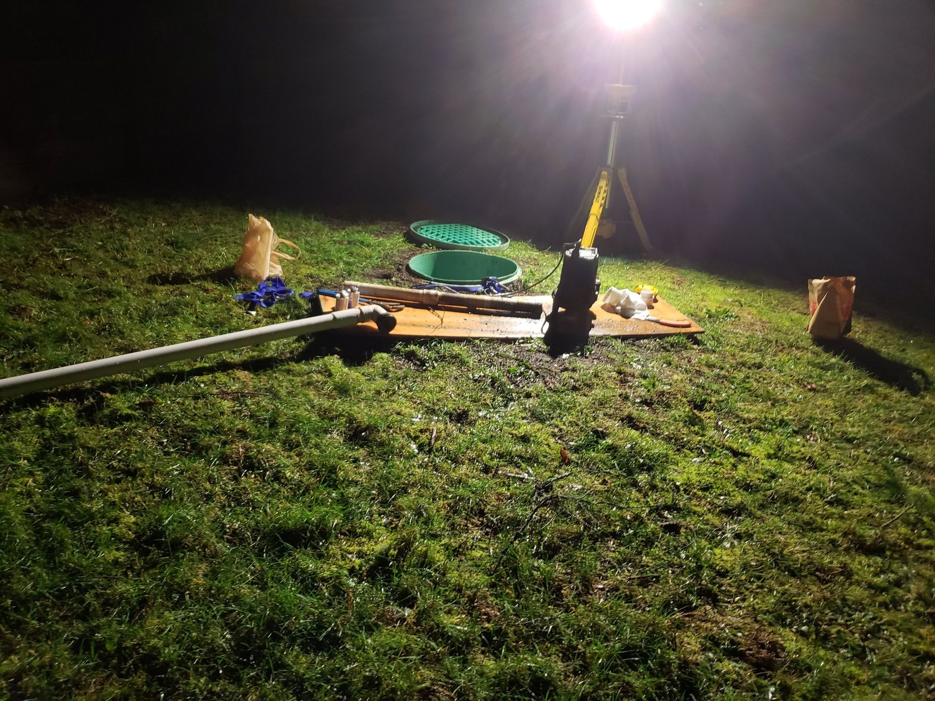 A tripod-mounted work light illuminates a green septic tank lid and maintenance equipment on a grassy lawn at night.