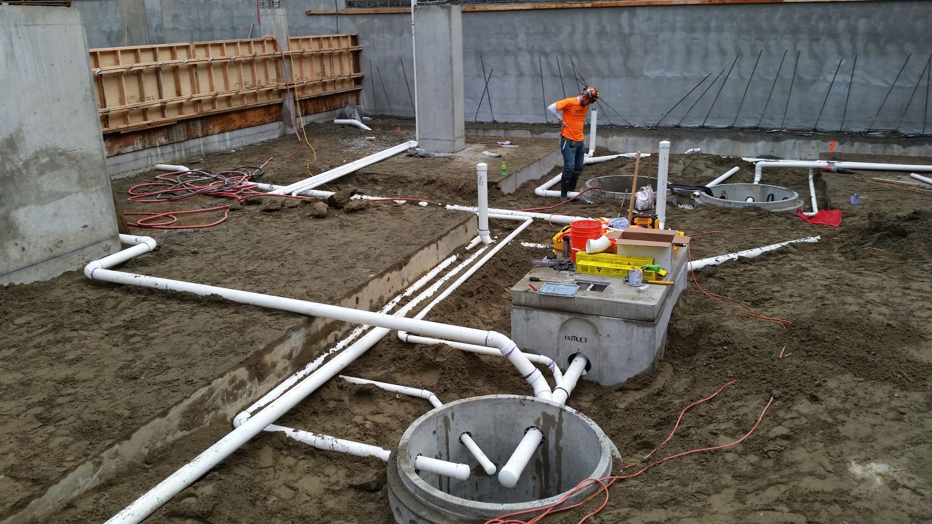 A worker in an orange shirt installs white PVC plumbing pipes on a dirt construction site foundation.