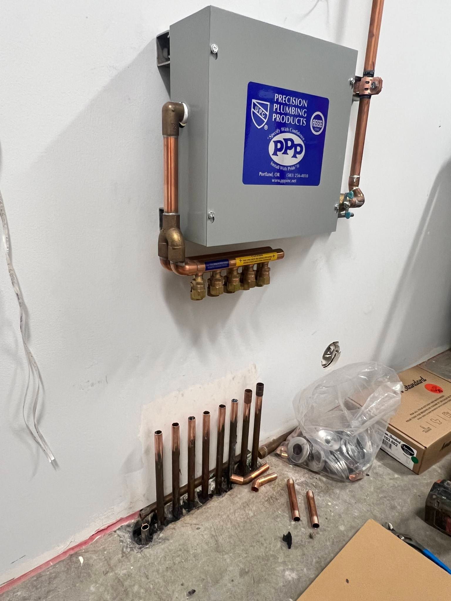 A water manifold and control box mounted on a white wall above a series of unfinished vertical copper pipes on the floor.