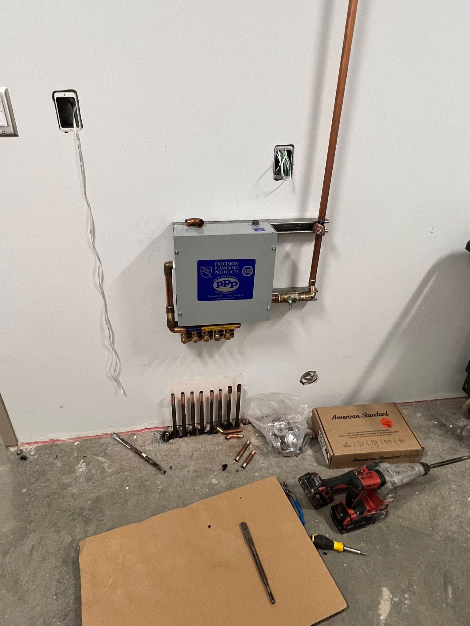 A mechanical box mounted on a wall with copper piping connected to a manifold below, amidst construction tools on a floor.