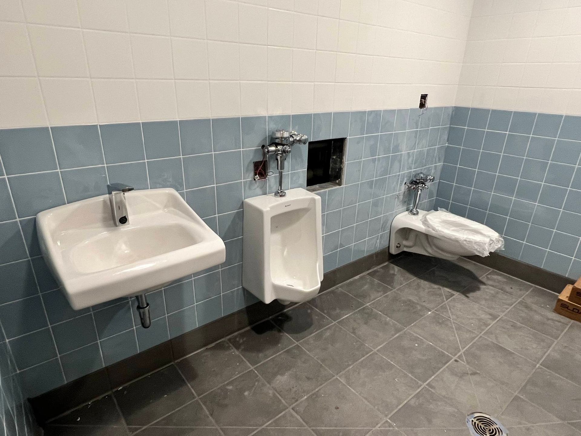 A bathroom with a white sink, a wall-mounted urinal, and a toilet seat covered in plastic against blue and white tiled walls.