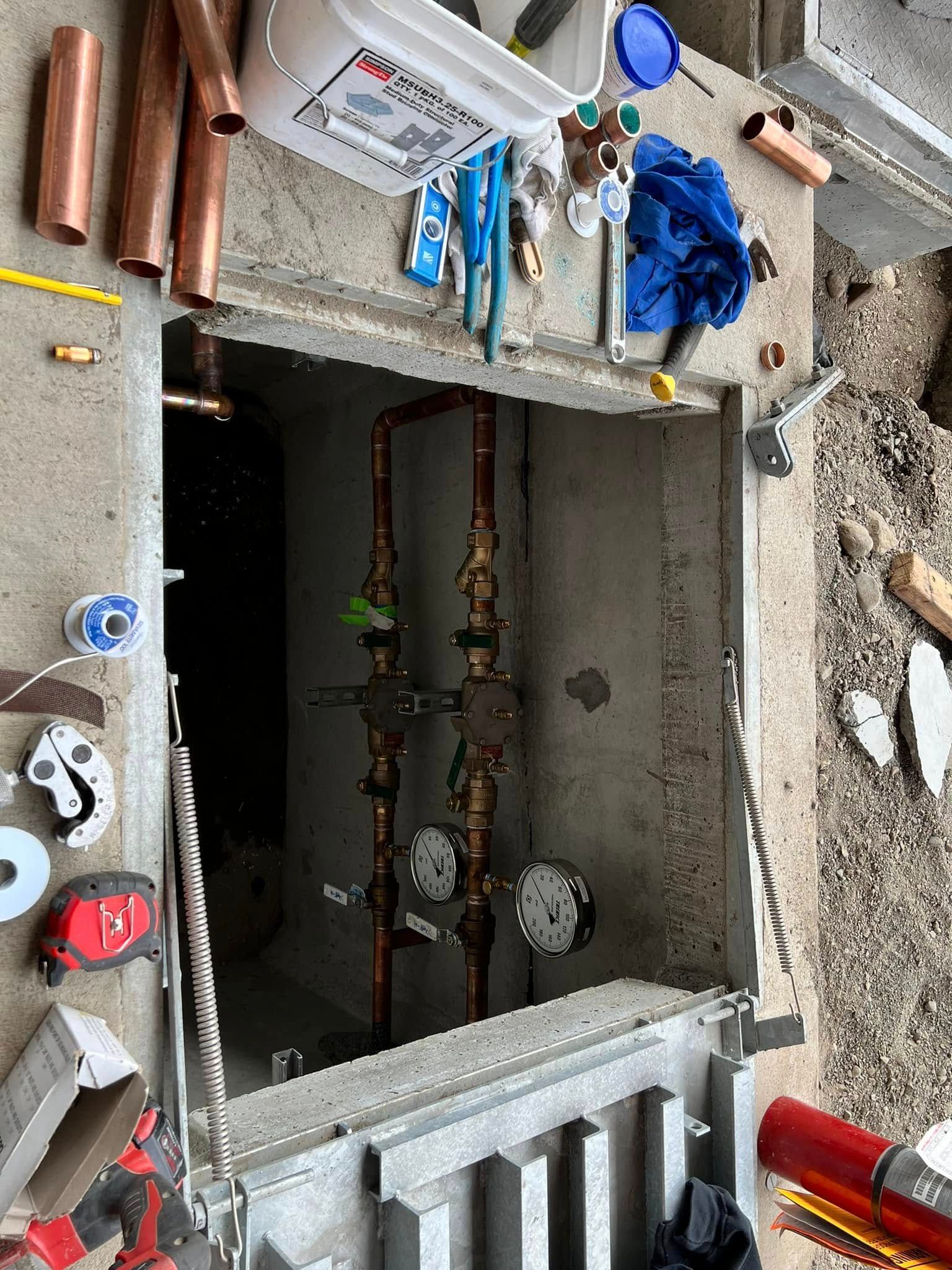 A concrete utility pit contains two vertical copper pipes with valves and a gauge. Tools and materials sit on the surface.