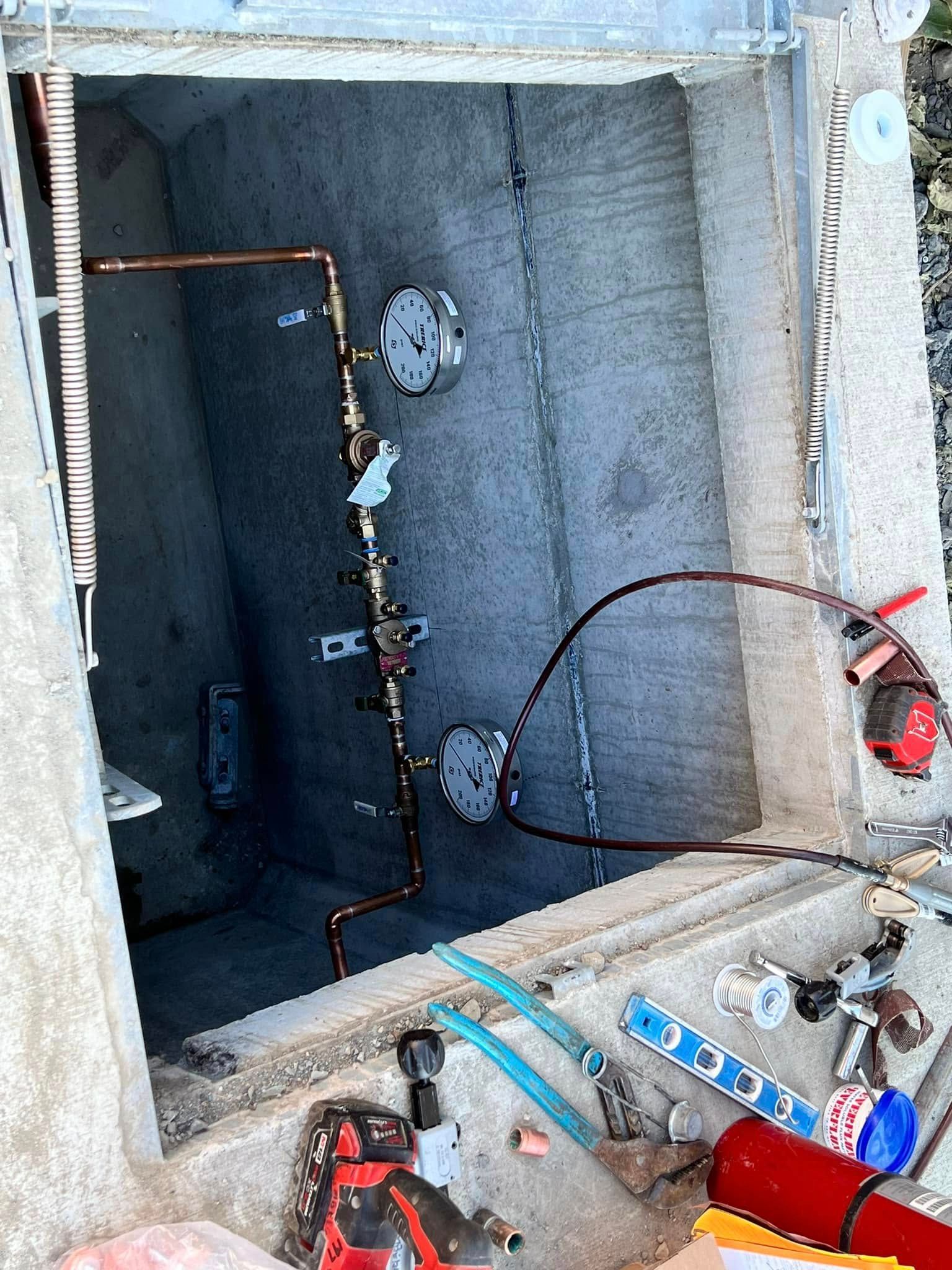 An open concrete utility pit containing copper piping with two circular gauges and various tools scattered on the edge.