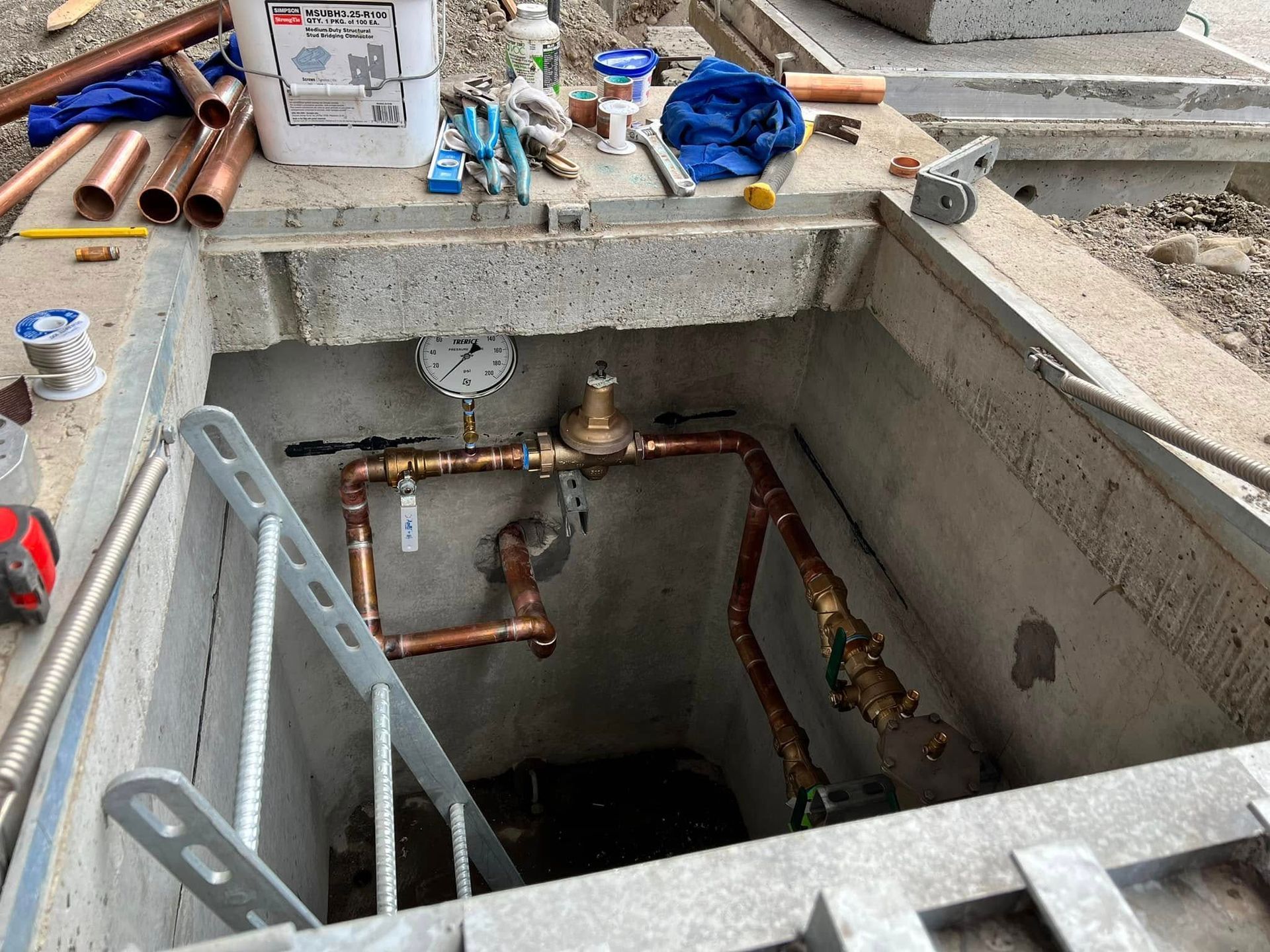 A concrete utility vault featuring a copper pipe water system with a gauge, pressure valve, and tools on the edge.