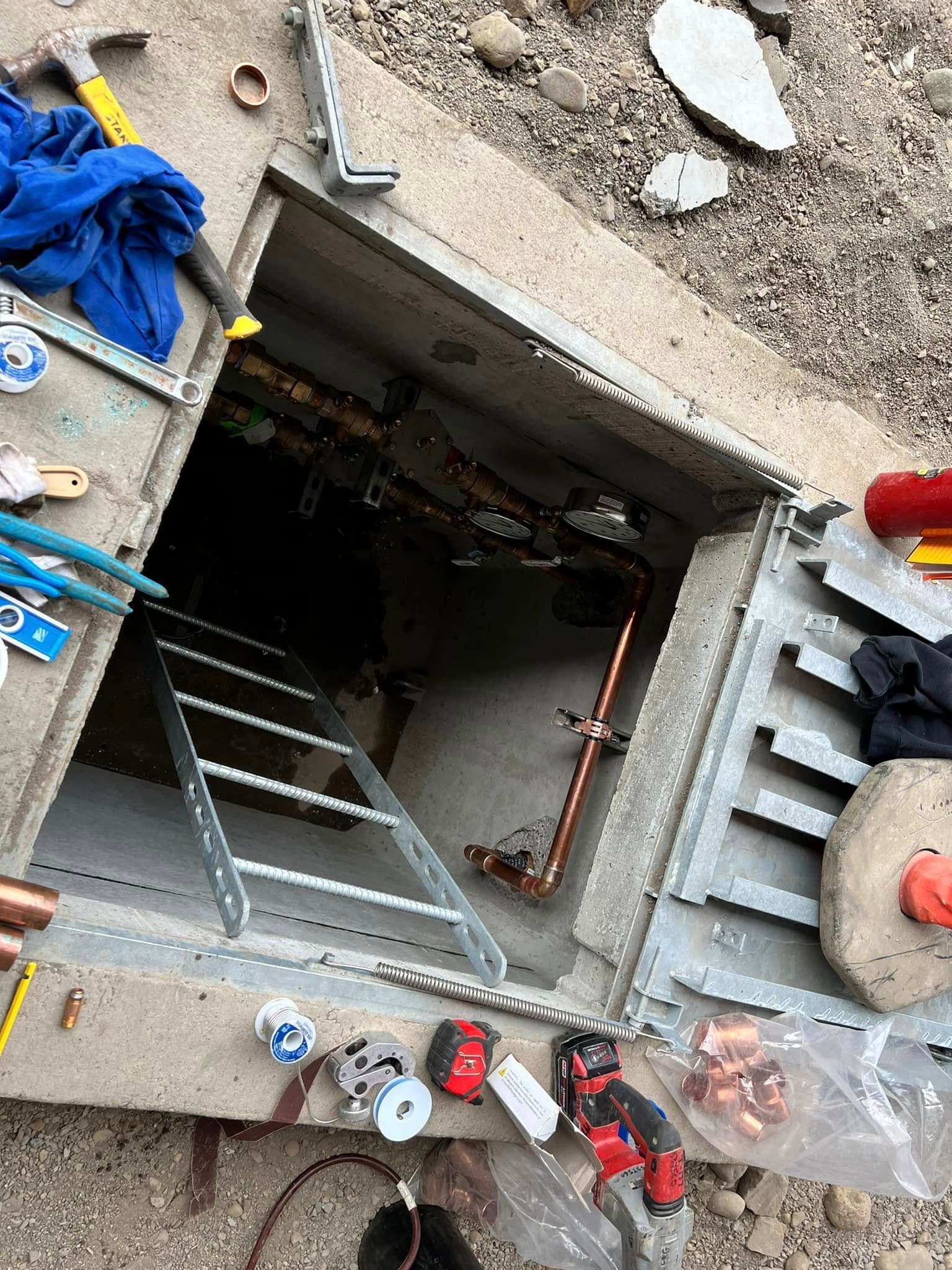 An open utility vault in the ground, showing copper plumbing, a metal ladder, and surrounding construction tools.