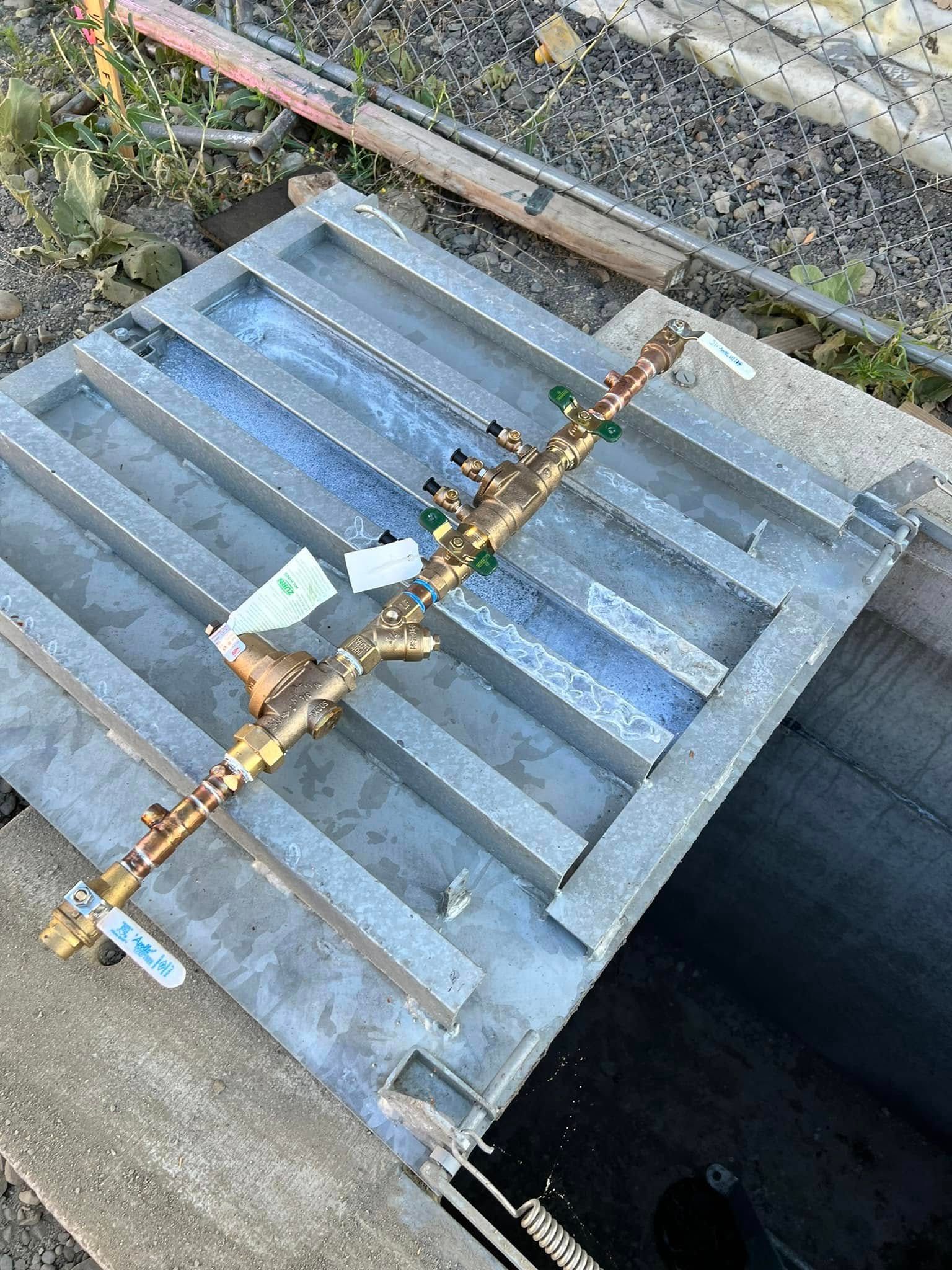 A brass water manifold assembly with multiple valves rests on a metal grate above a utility pit.