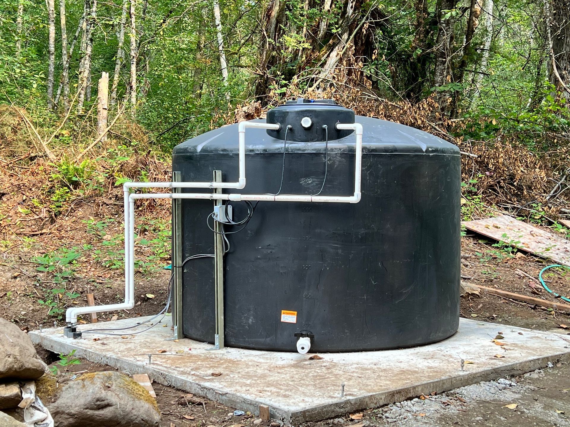 A black water storage tank on a concrete pad in a wooded area, equipped with white PVC piping.
