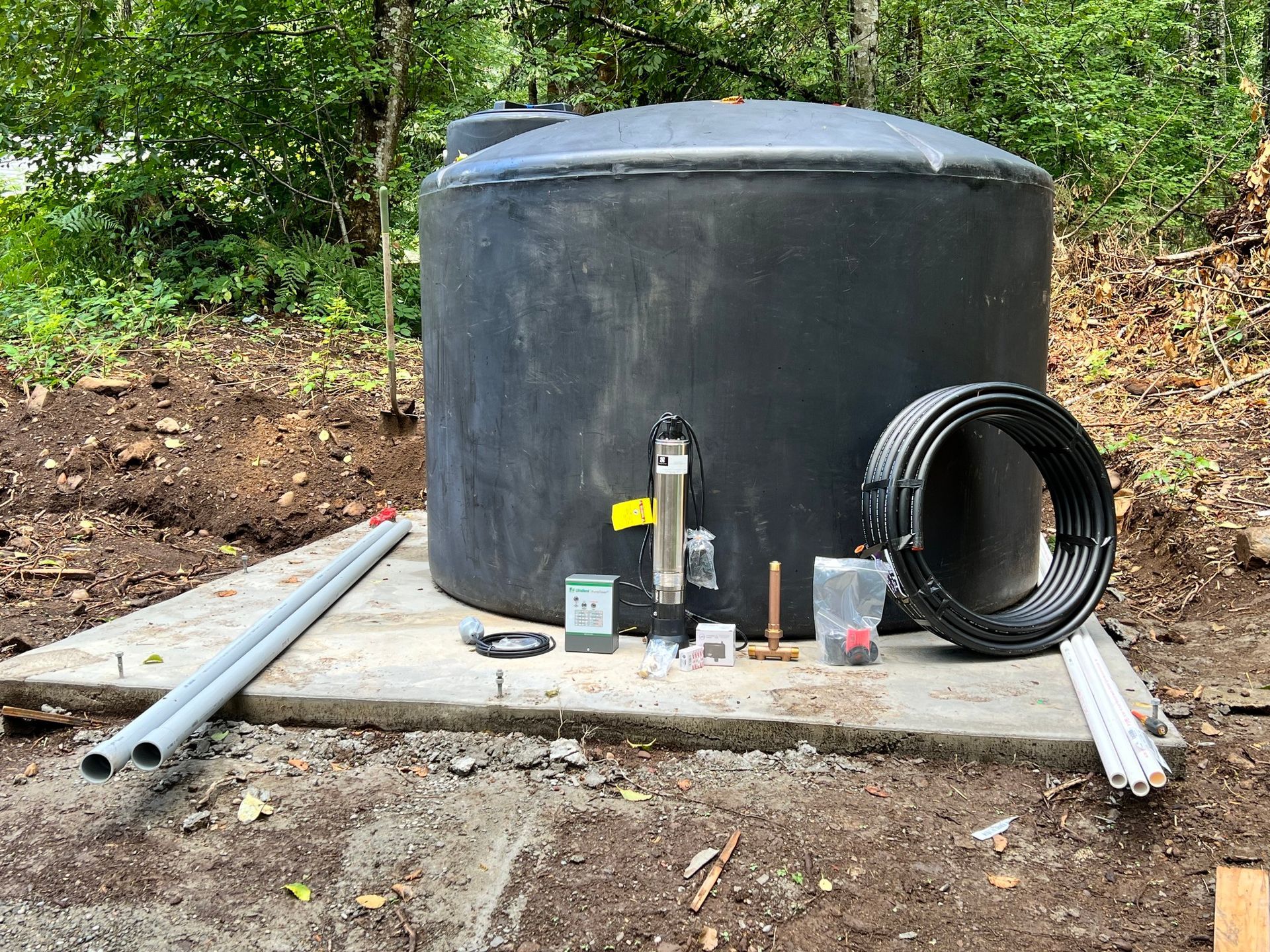 A large black plastic water tank sits on a concrete pad in a wooded area with plumbing supplies and piping arranged around it.