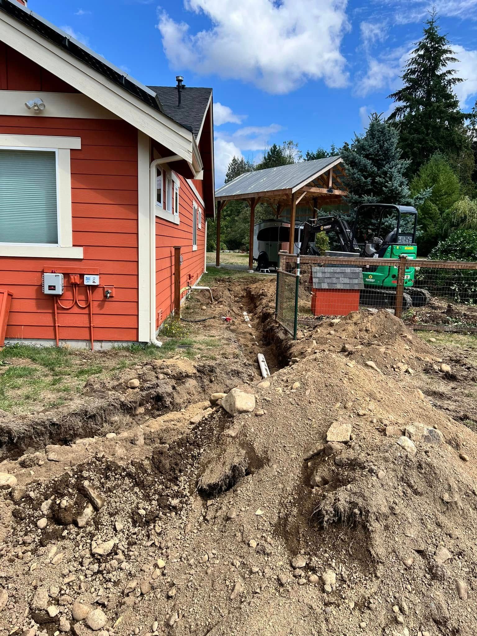 A side view of a red house with a trench dug in the dirt yard leading toward a small wooden structure with a backhoe.
