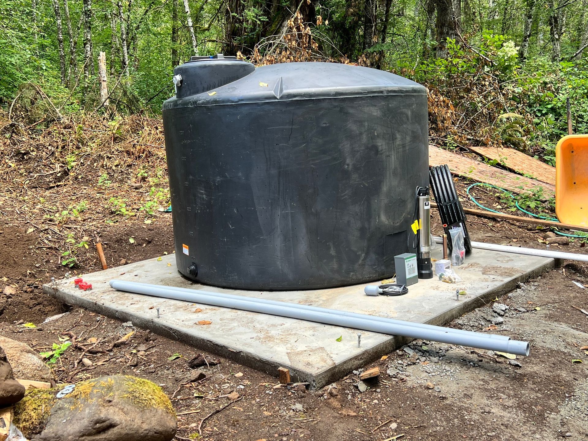 A large black cylindrical water storage tank sits on a concrete pad outdoors in a wooded area with plumbing components.