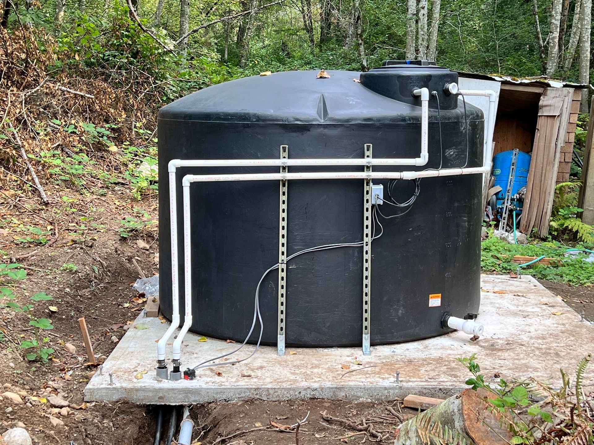 A large, black circular water tank sits on a concrete pad outdoors, featuring white PVC piping and metal support rails.