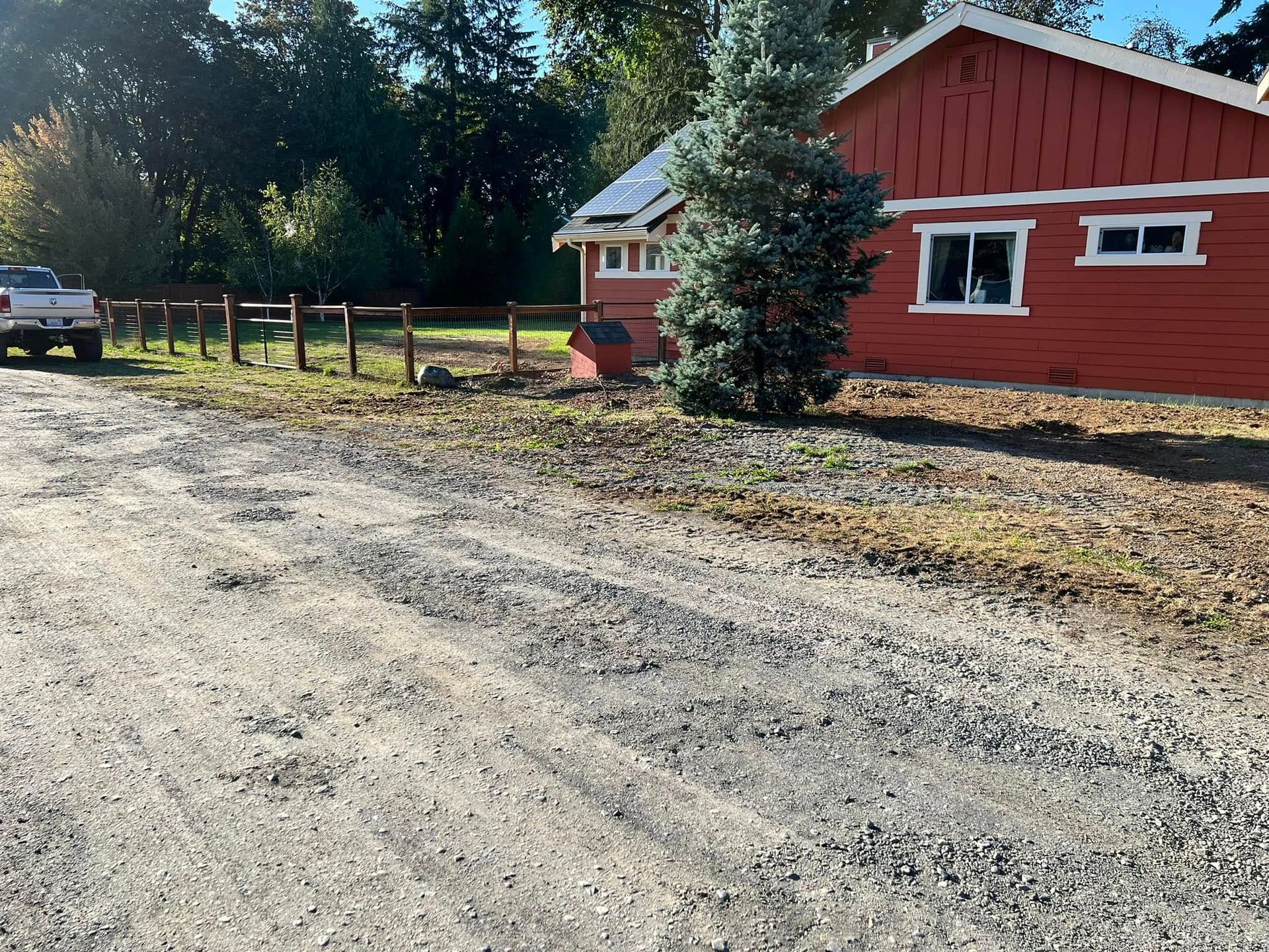 A gravel driveway leads to a red, wood-sided house next to a fence and evergreen tree, with a white truck parked nearby.