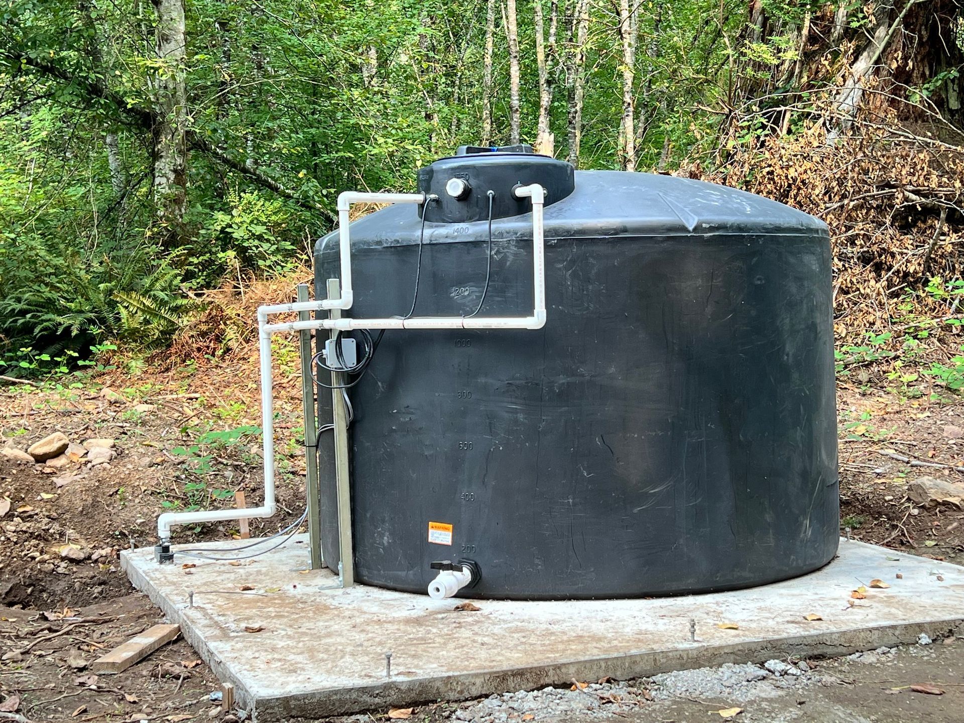 A large black water storage tank sitting on a concrete pad in a wooded outdoor area, fitted with white PVC piping.