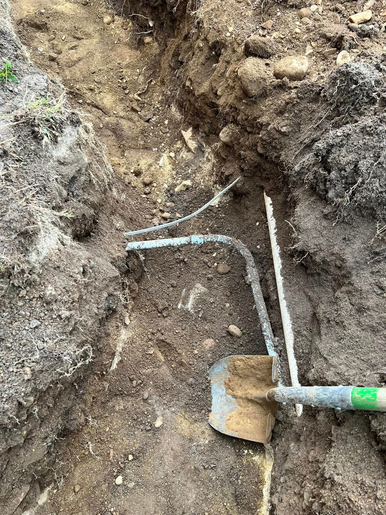 A shovel rests in a dirt trench next to several exposed, L-shaped white PVC pipes.