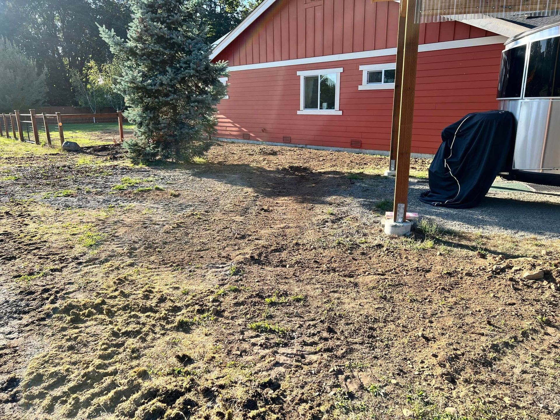 A red farmhouse exterior with a muddy yard, a large pine tree, and a covered travel trailer in the foreground.
