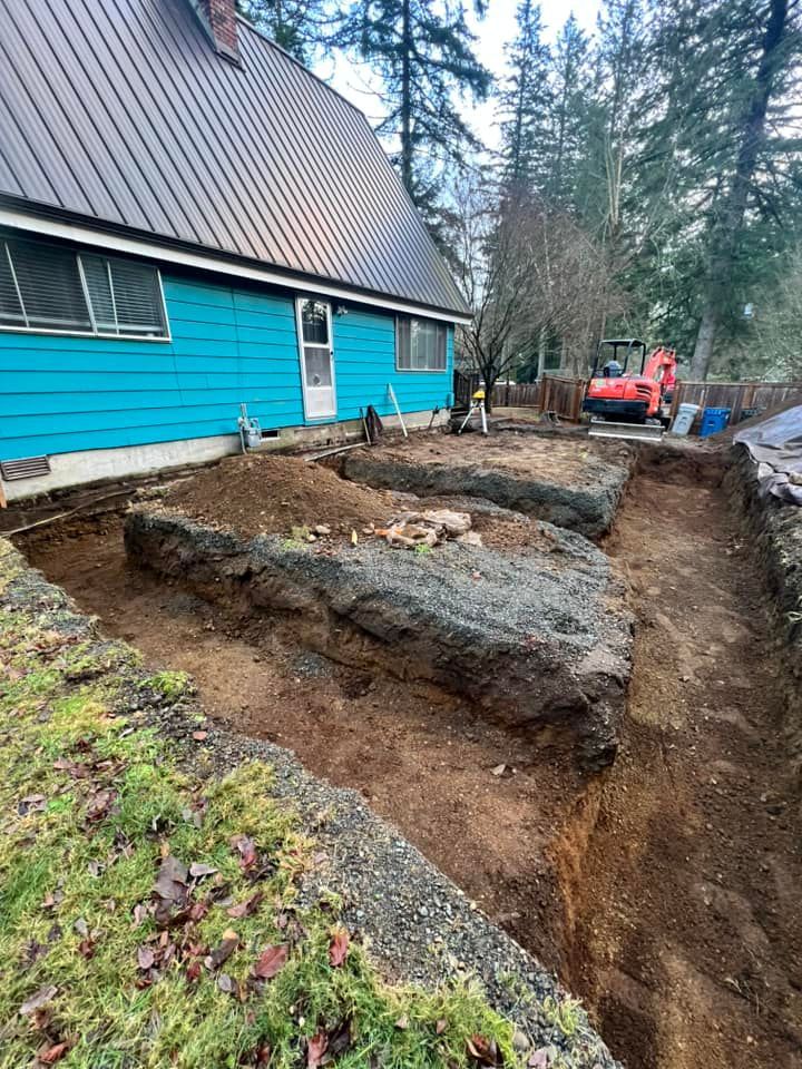 A teal house with a brown roof sits beside a backyard construction site with excavated dirt trenches and a small excavator.
