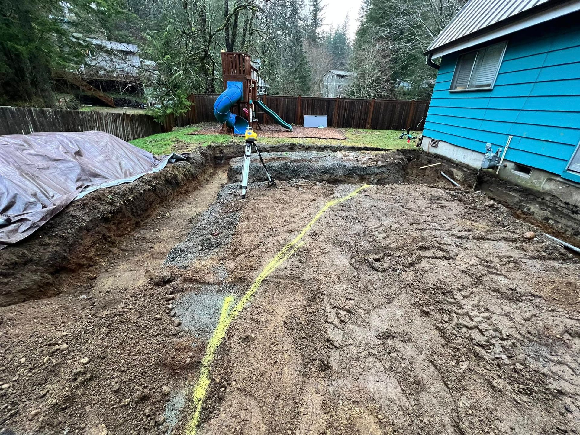 An excavation site with a tripod laser level, a trench, and a blue house under construction in a wooded backyard.