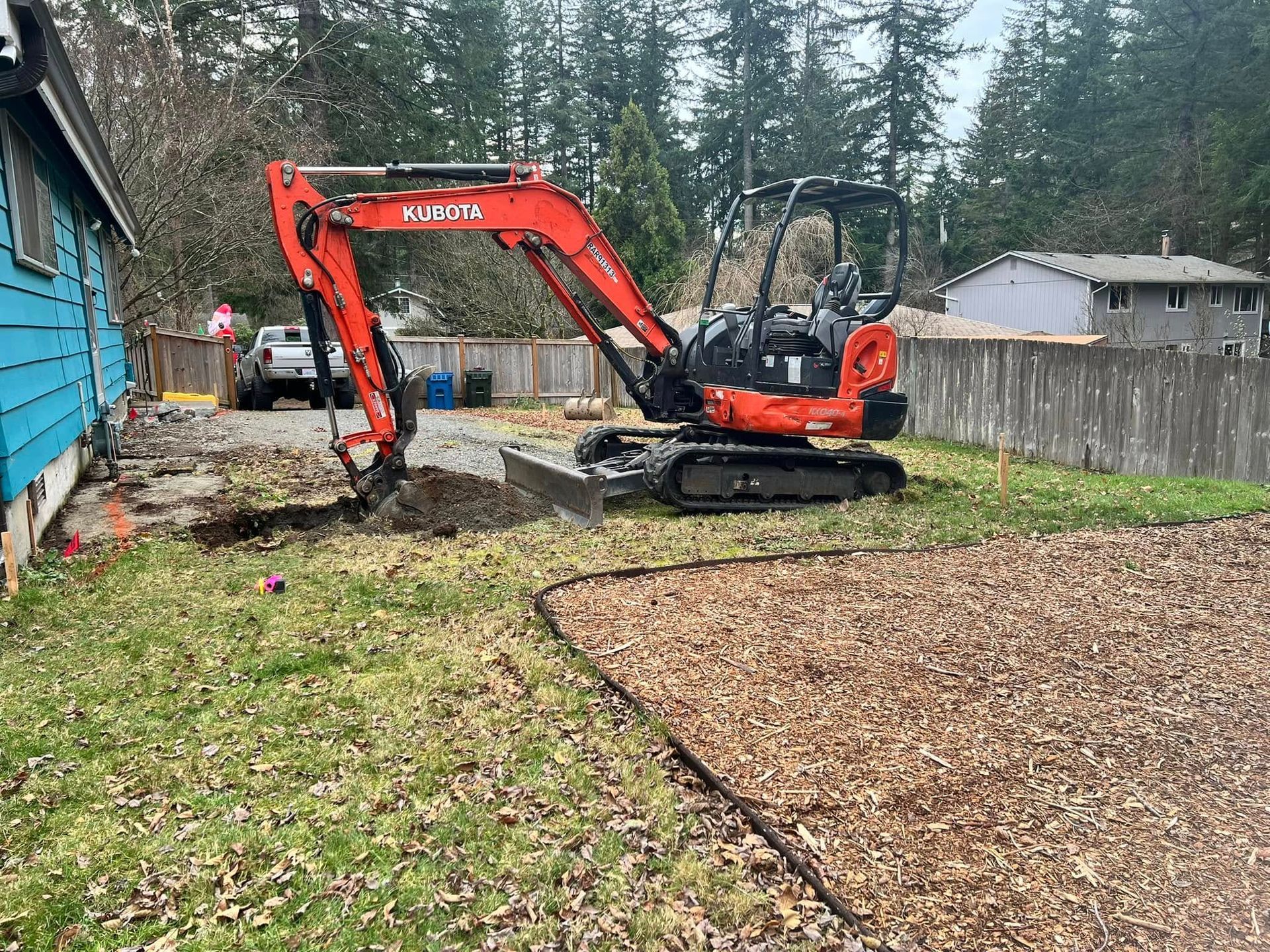 An orange Kubota excavator parked in a residential yard near the side of a blue house, digging into the grass.
