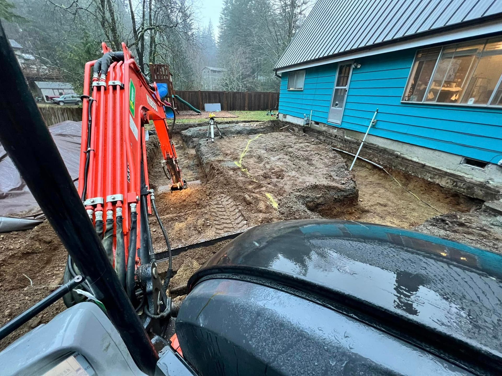 An orange excavator operates near the side of a blue house, digging a trench in a muddy yard.