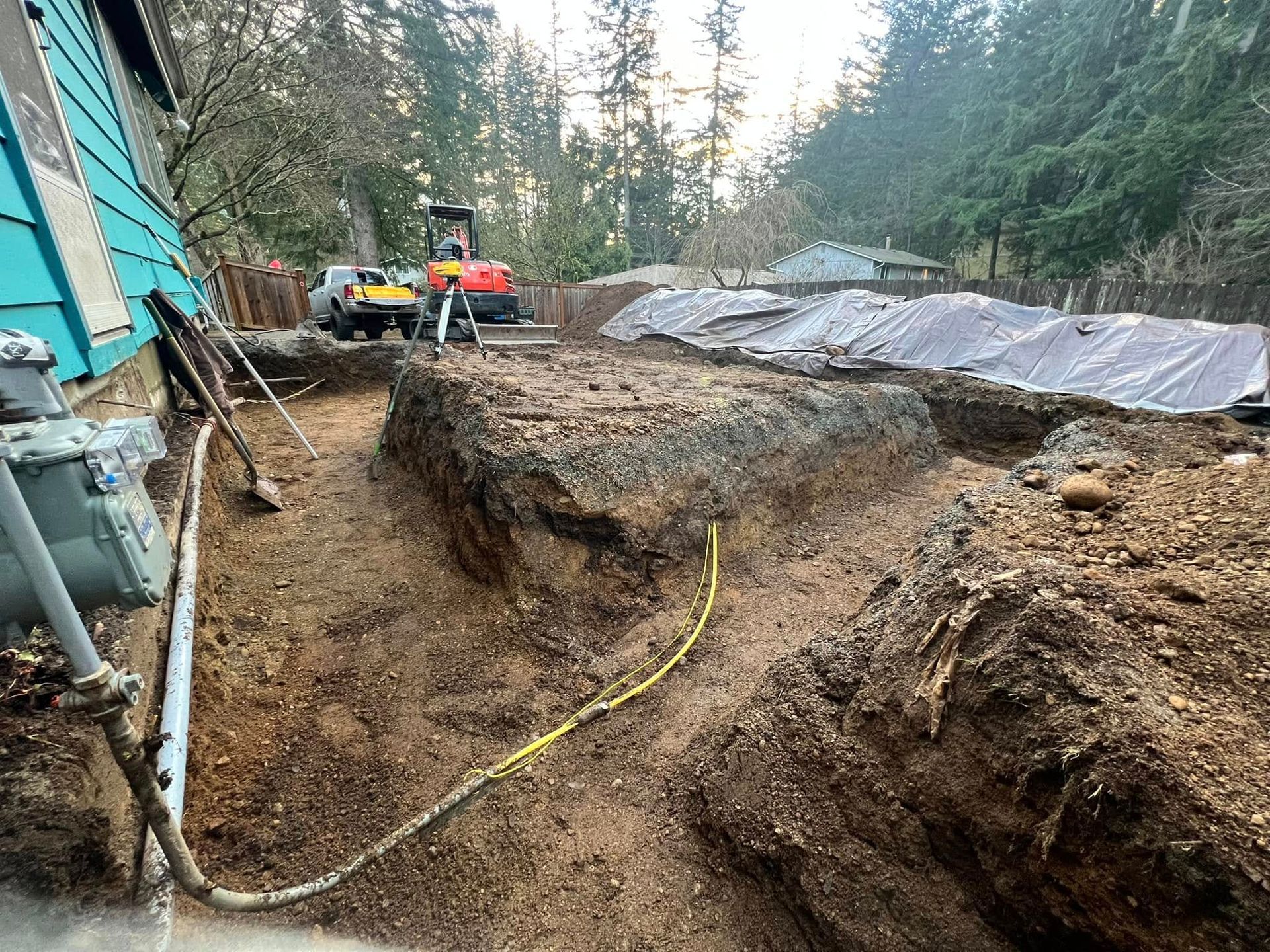 Excavated trench and foundation work next to a house, with construction equipment and dirt piles in a wooded setting.