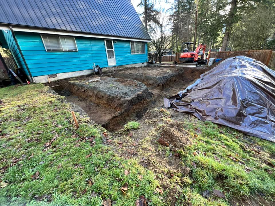 An excavated foundation area next to a bright blue house, with a pile of dirt covered by a plastic tarp in the yard.