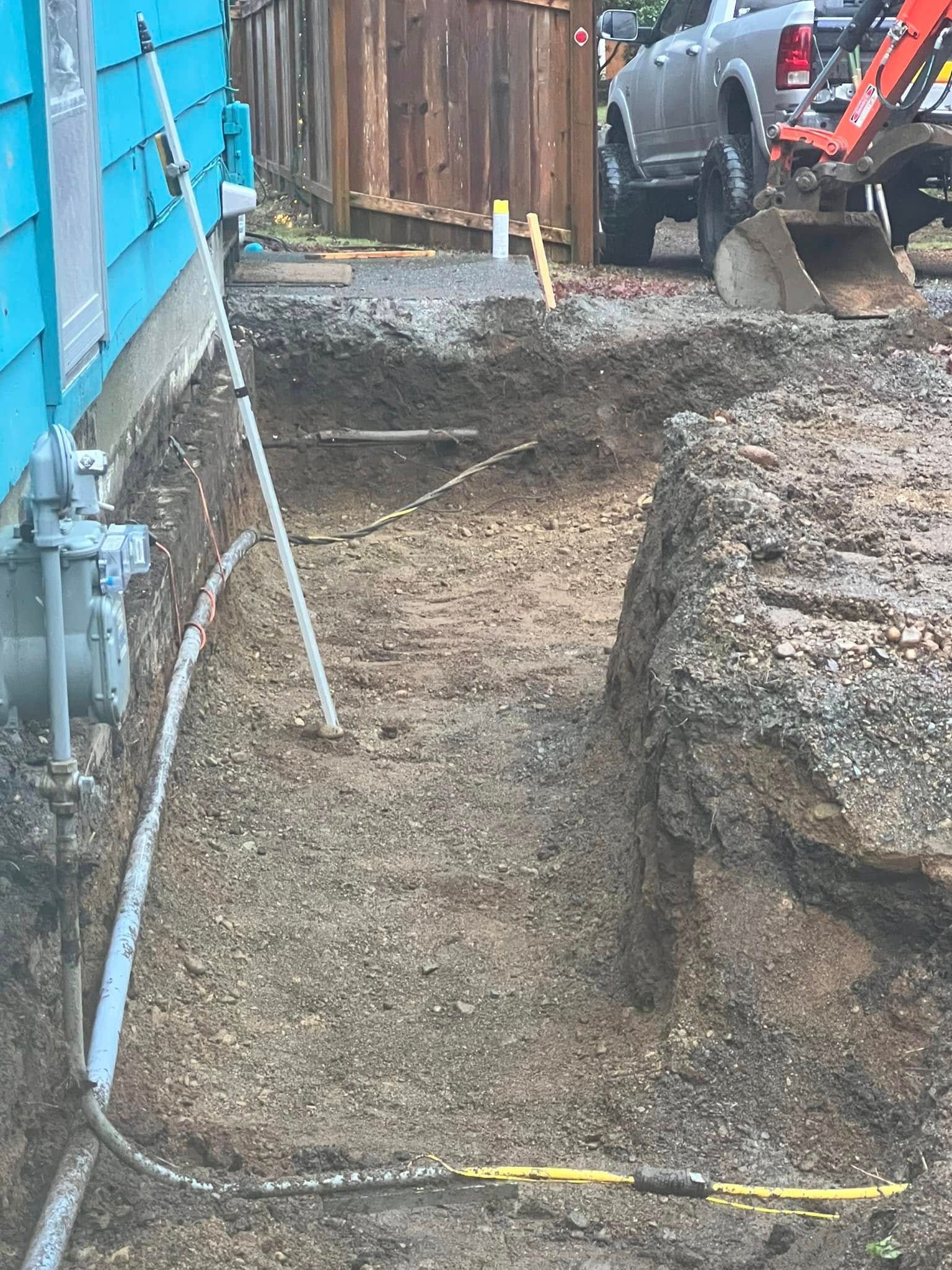 A trench dug alongside the foundation of a blue-sided house with construction equipment in the background.
