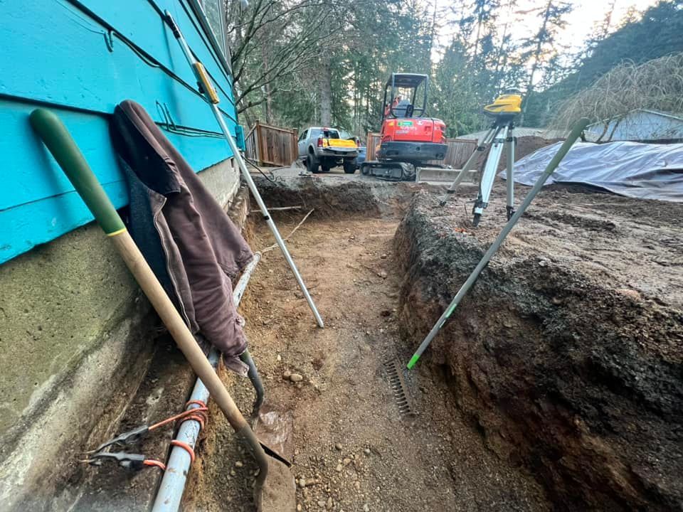 A trench runs along a blue building foundation at a construction site with an excavator and surveying tripod in the back.