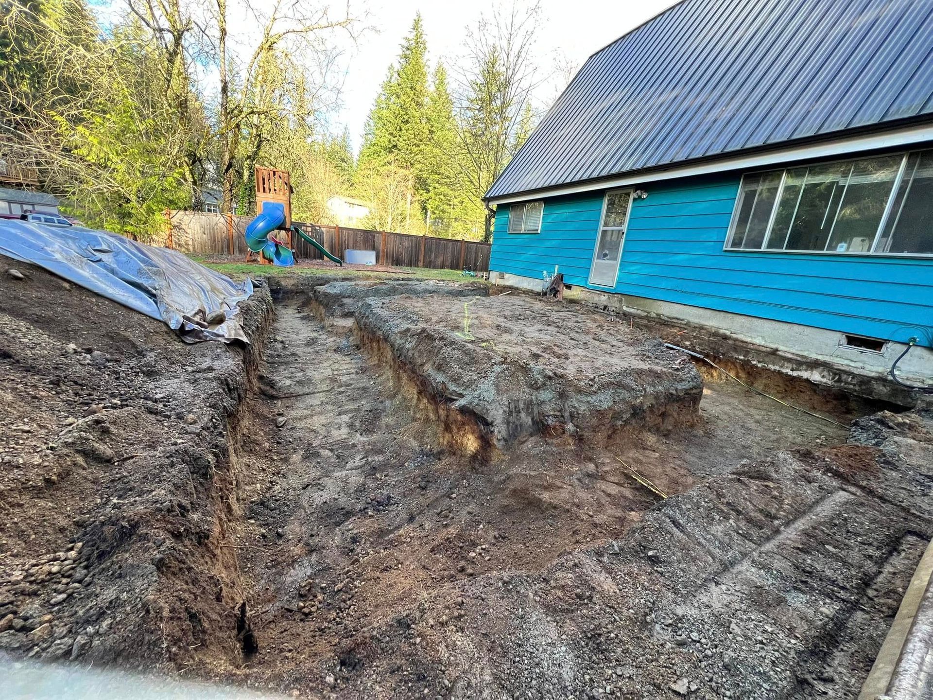 An excavation site with a dirt trench beside a blue house with a metal roof and a play set in the background.