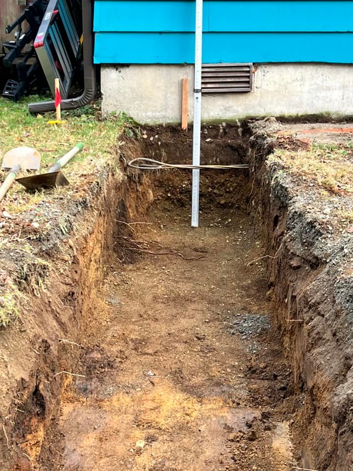 A deep, narrow trench dug into the ground in front of a house with blue siding and a white vertical pipe.