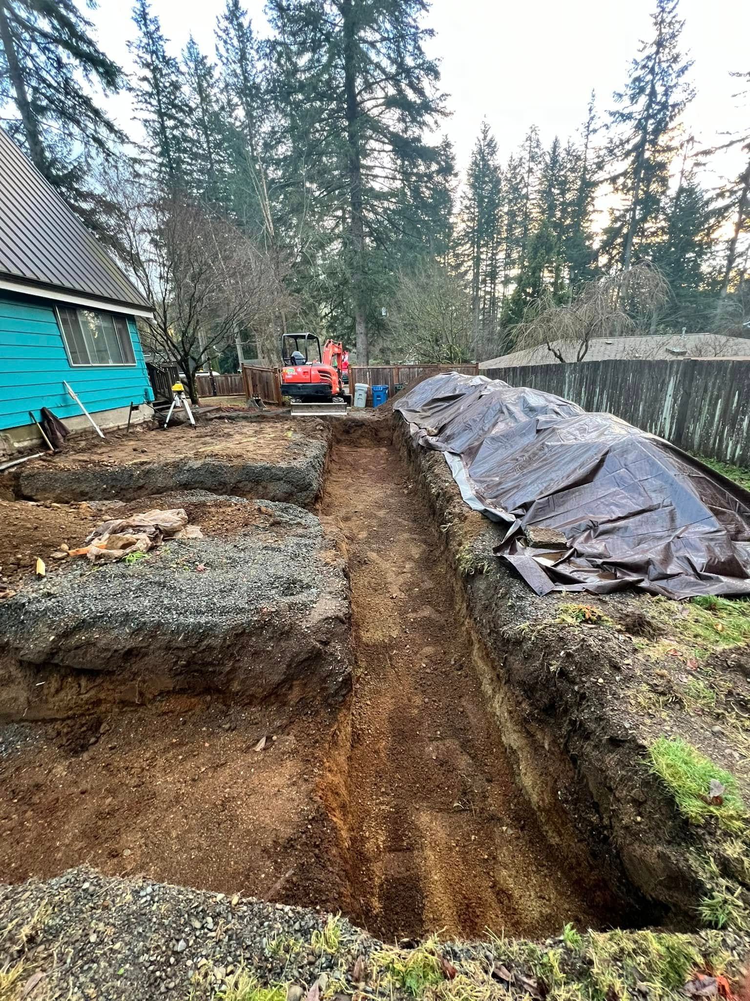 A construction site with a deep trench in the center, a blue-sided house on the left, and a covered soil pile on the right.