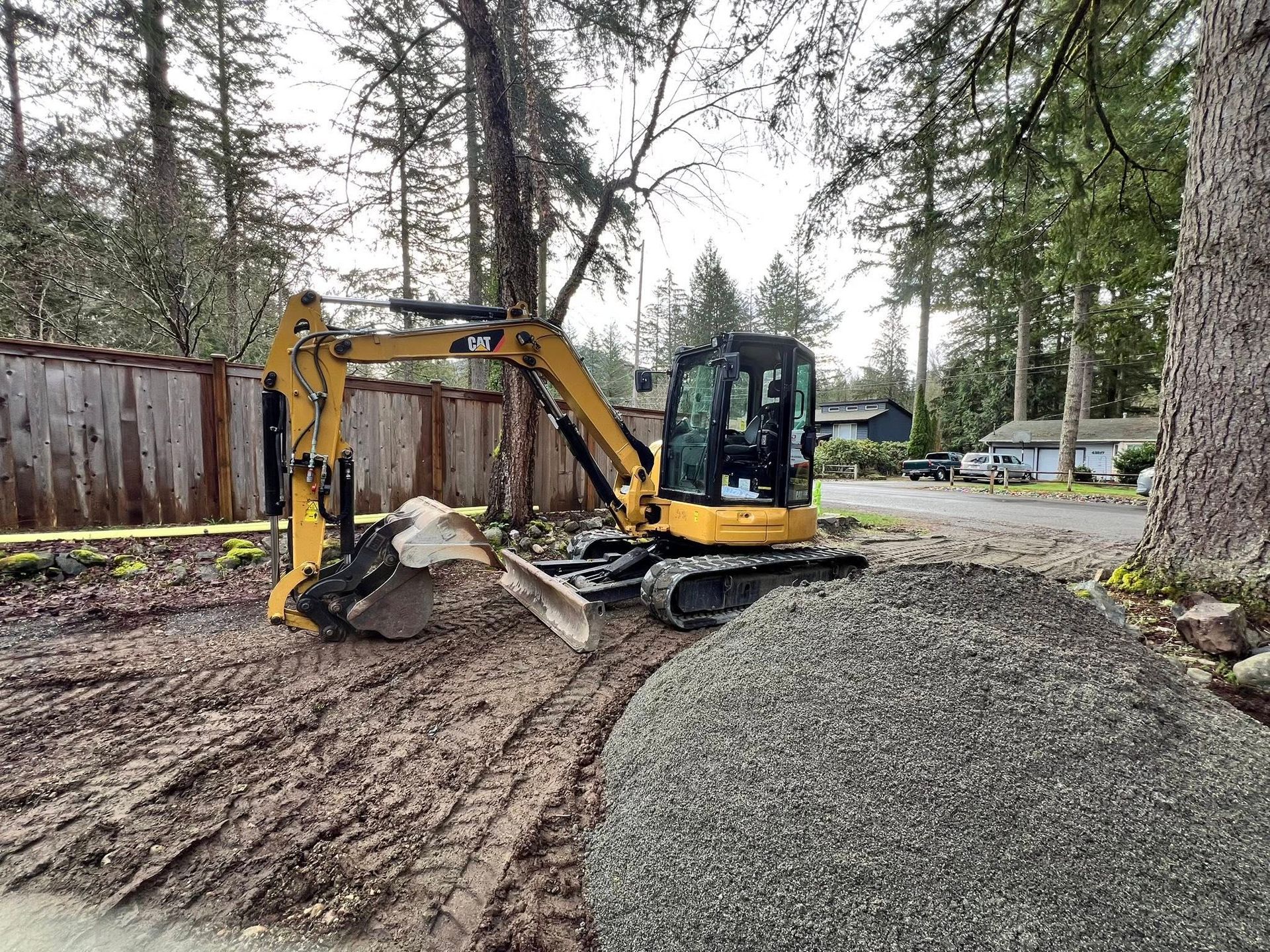 Yellow Caterpillar excavator parked on muddy ground next to a pile of gravel in a wooded residential area.